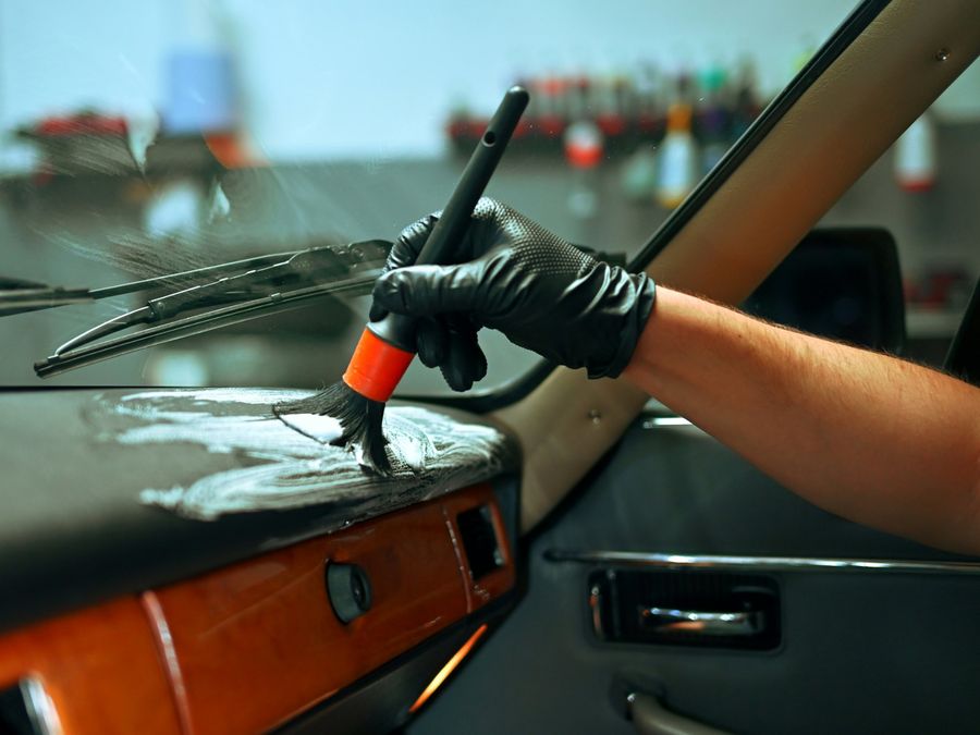 Gloved hand working foam cleaner into a car dashboard with a detailing brush